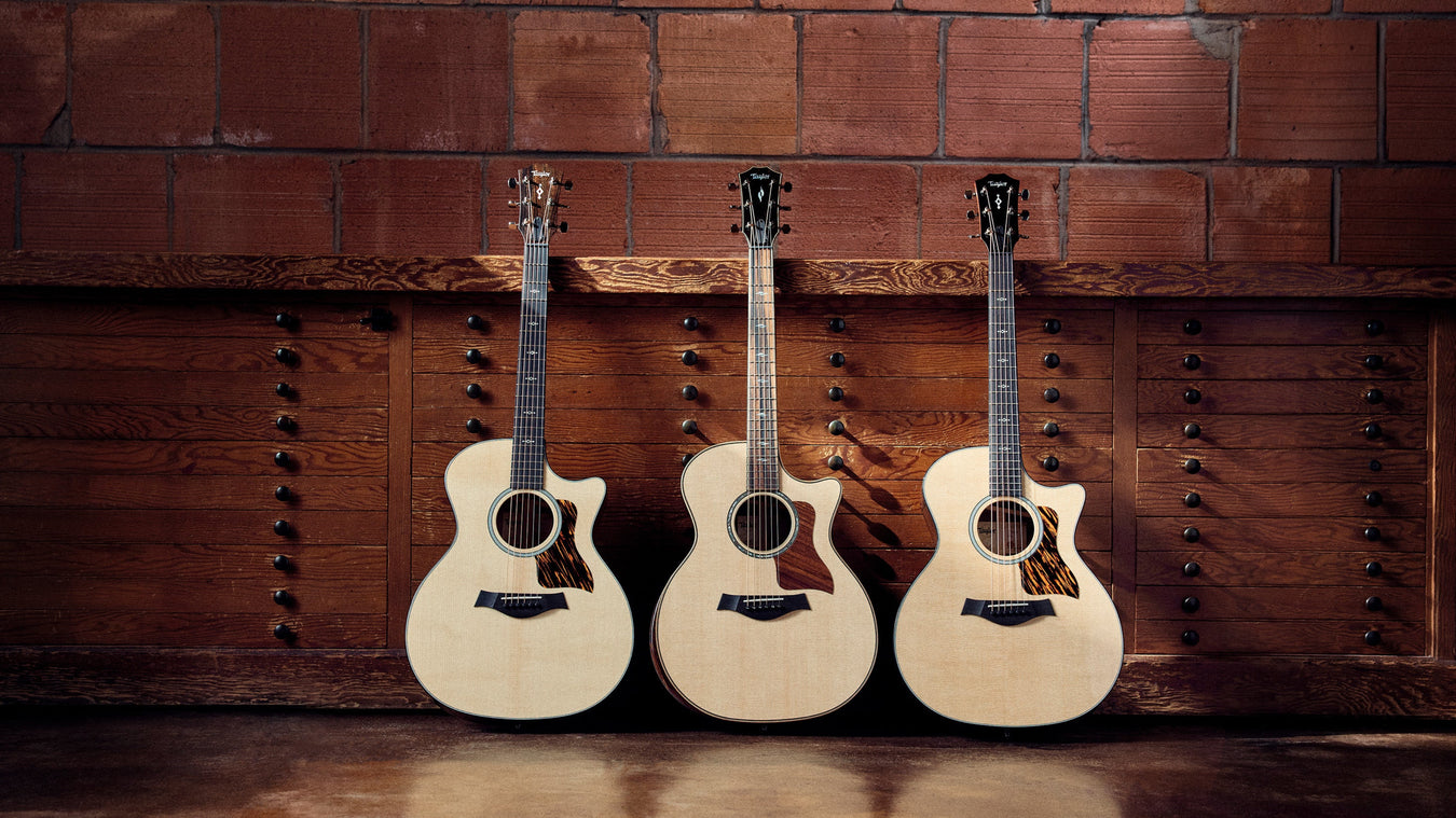 Three acoustic guitars on a brown concrete floor, leaned up against a wooden drawer and a brick wall.